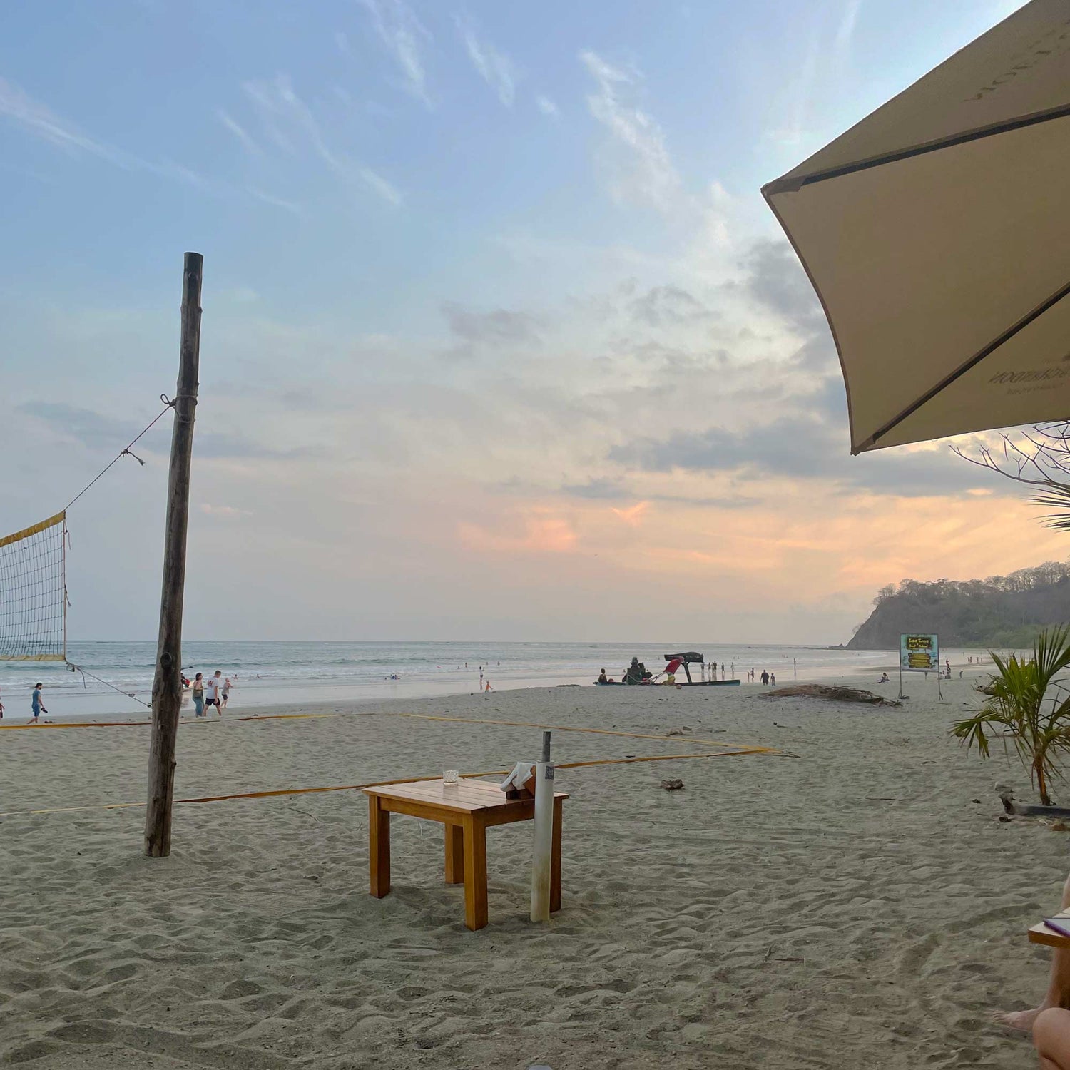 A woman sits at a table on the beach with a volleyball net