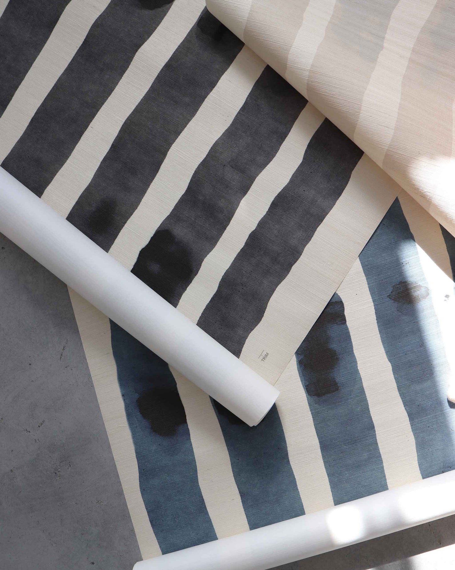 Three overlapping rolls of Custom Grasscloth, featuring irregular hand-painted stripes in black, blue, and beige, are displayed on a gray surface, echoing the unique look of bespoke grasscloth wallpaper.