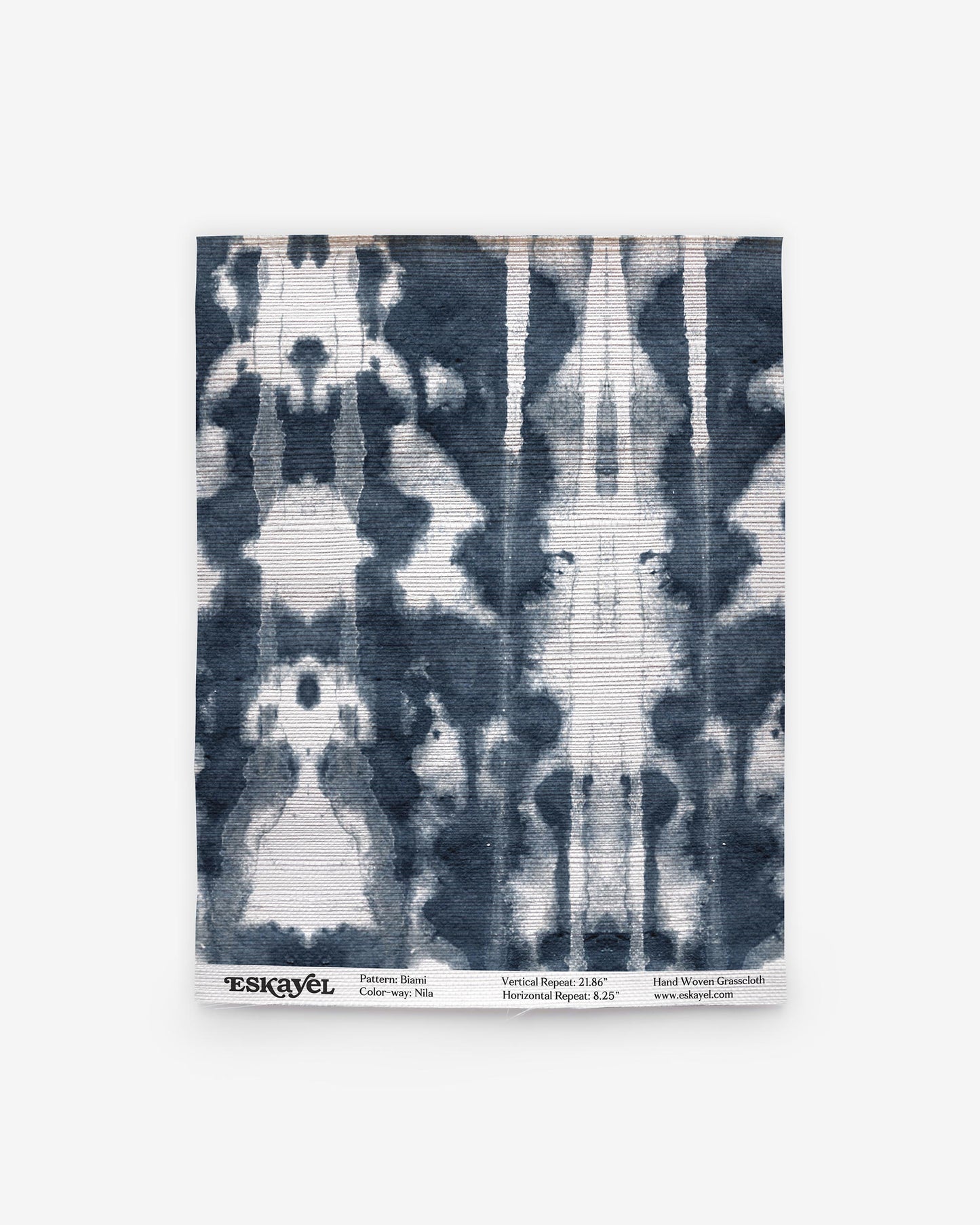 Rectangular fabric sample featuring a symmetrical indigo and white tie-dye pattern, reminiscent of blue-on-white woven textiles. Text at the bottom displays the brand name Eskayel along with details about the pattern, color (Biami Grasscloth||Nila), and website information.