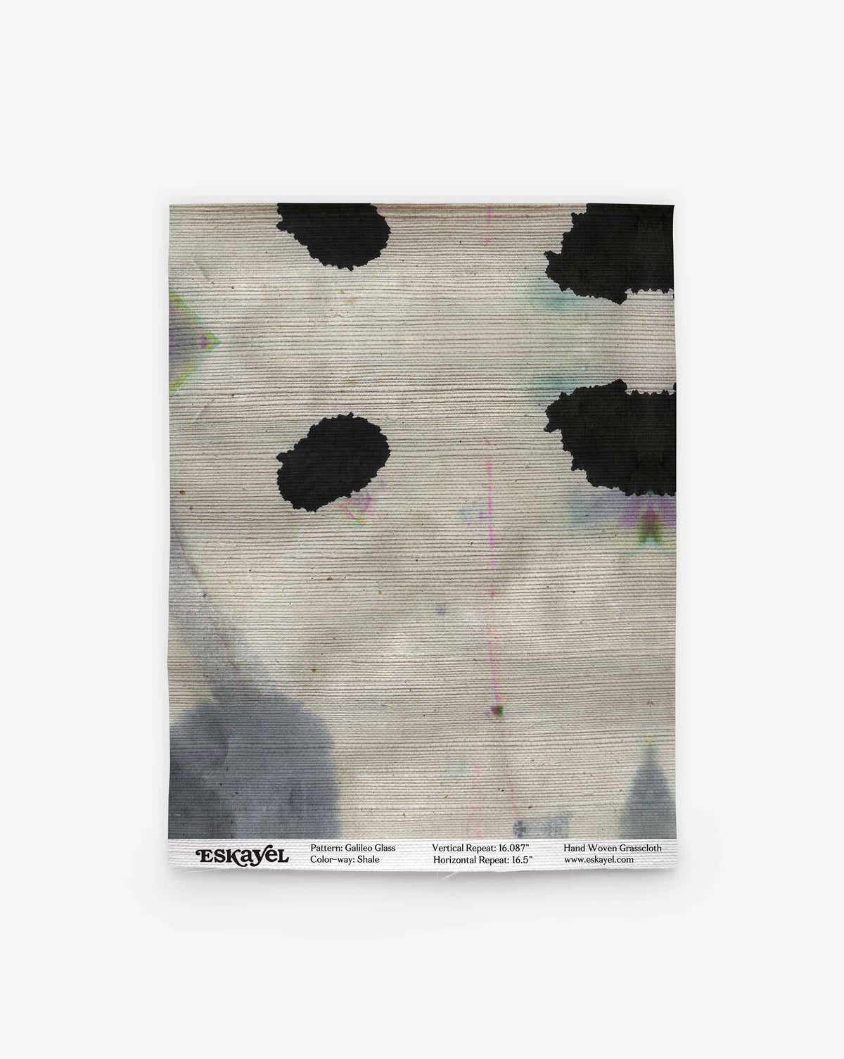 A folded textile featuring the Galileo Glass Grasscloth in the Shale colorway, showcasing an abstract black spot pattern on a light gray background, displayed against a white surface—evoking high-end wallpaper aesthetics. Text at the bottom notes the pattern, color (Shale), size, and brand details.