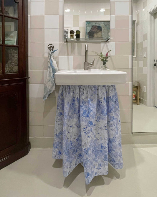 A white bathroom sink with a skirt made from Crochet Lace Fabric||Fresco Blue, paired with a hand towel, small flower vase, and tiled walls.