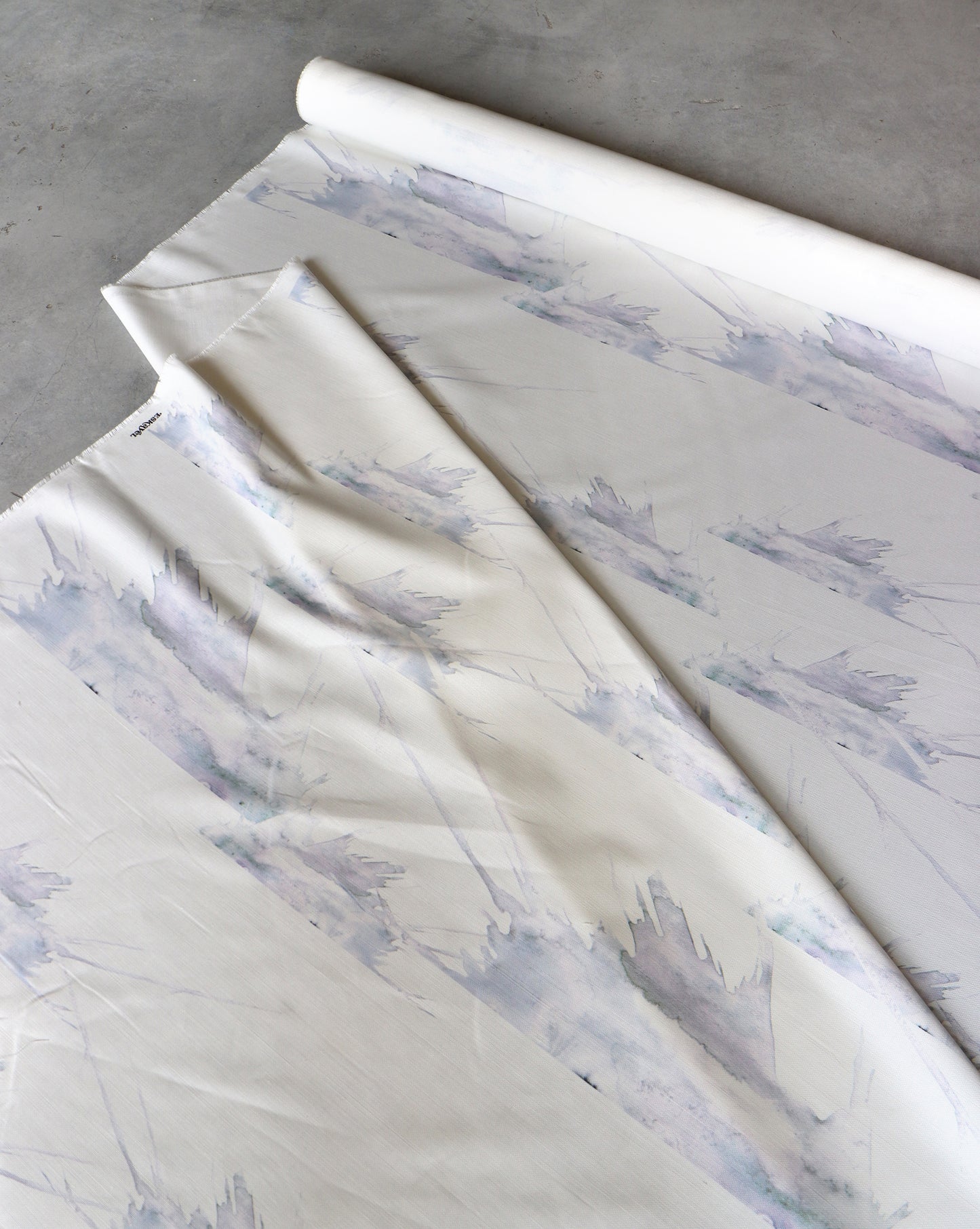 A roll of "Love in the Middle Fabric ||Ice," a light-colored linen/cotton blend with a watercolor-style pattern, 54 inches wide and 14 yards long, is partially unrolled on a surface.
