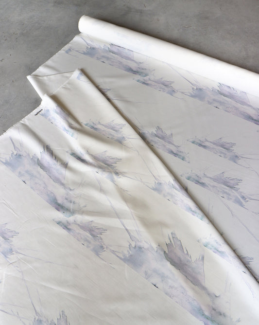 A roll of "Love in the Middle Fabric ||Ice," a light-colored linen/cotton blend with a watercolor-style pattern, 54 inches wide and 14 yards long, is partially unrolled on a surface.
