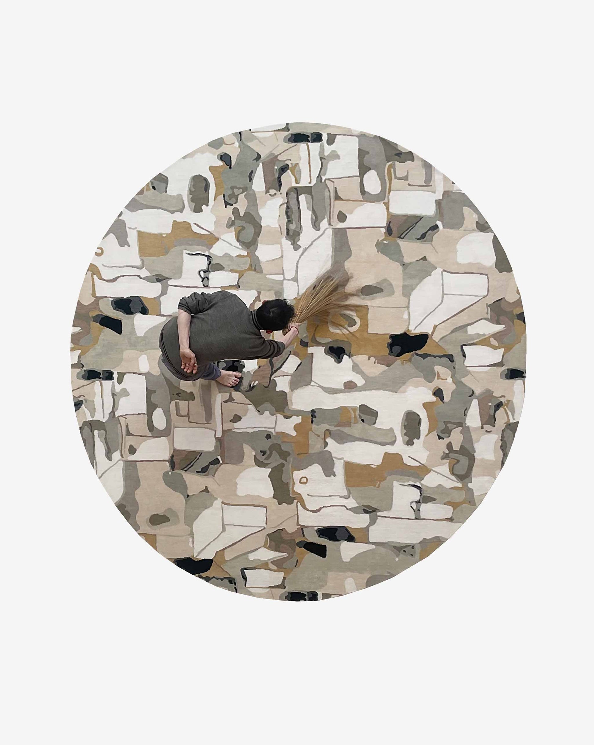 A person seen from above uses a broom on the Medina Hand Knotted Rug||Blanca CUSTOM, a circular, abstract-patterned handmade rug in neutral colors.