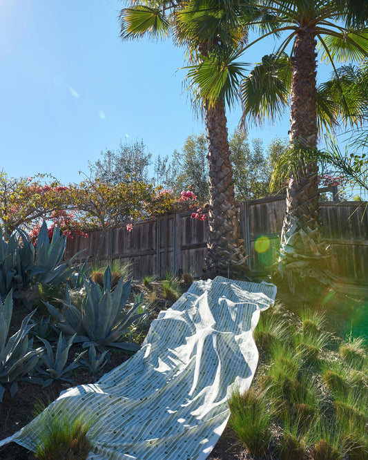 A blanket made of Bamboo Stripe Performance Fabric is laying on the ground in front of a palm tree