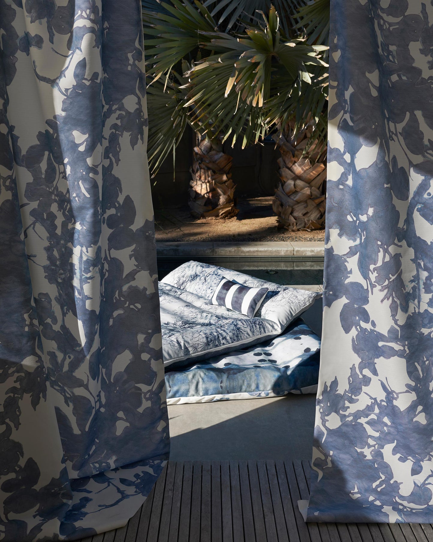 Outdoor relaxation area with Up for Anything Performance Fabric||Cerulean patterned curtains, cushions, and a throw on a wooden deck, surrounded by palm trees.