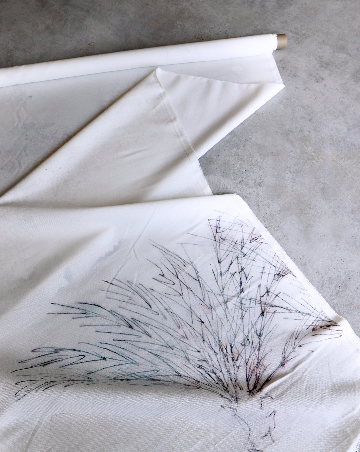 A roll of Palmeraie Fabric ||Duomo, measuring 54 inches wide and featuring a minimalistic tree sketch, is partially unrolled on a concrete surface, showcasing its elegant details. This linen cotton fabric is perfect for various creative projects and sold by the yard.