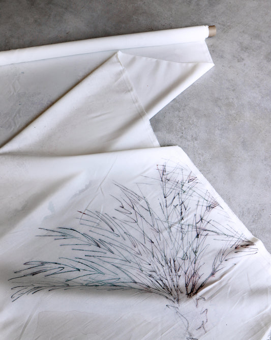 A roll of Palmeraie Fabric ||Duomo, measuring 54 inches wide and featuring a minimalistic tree sketch, is partially unrolled on a concrete surface, showcasing its elegant details. This linen cotton fabric is perfect for various creative projects and sold by the yard.