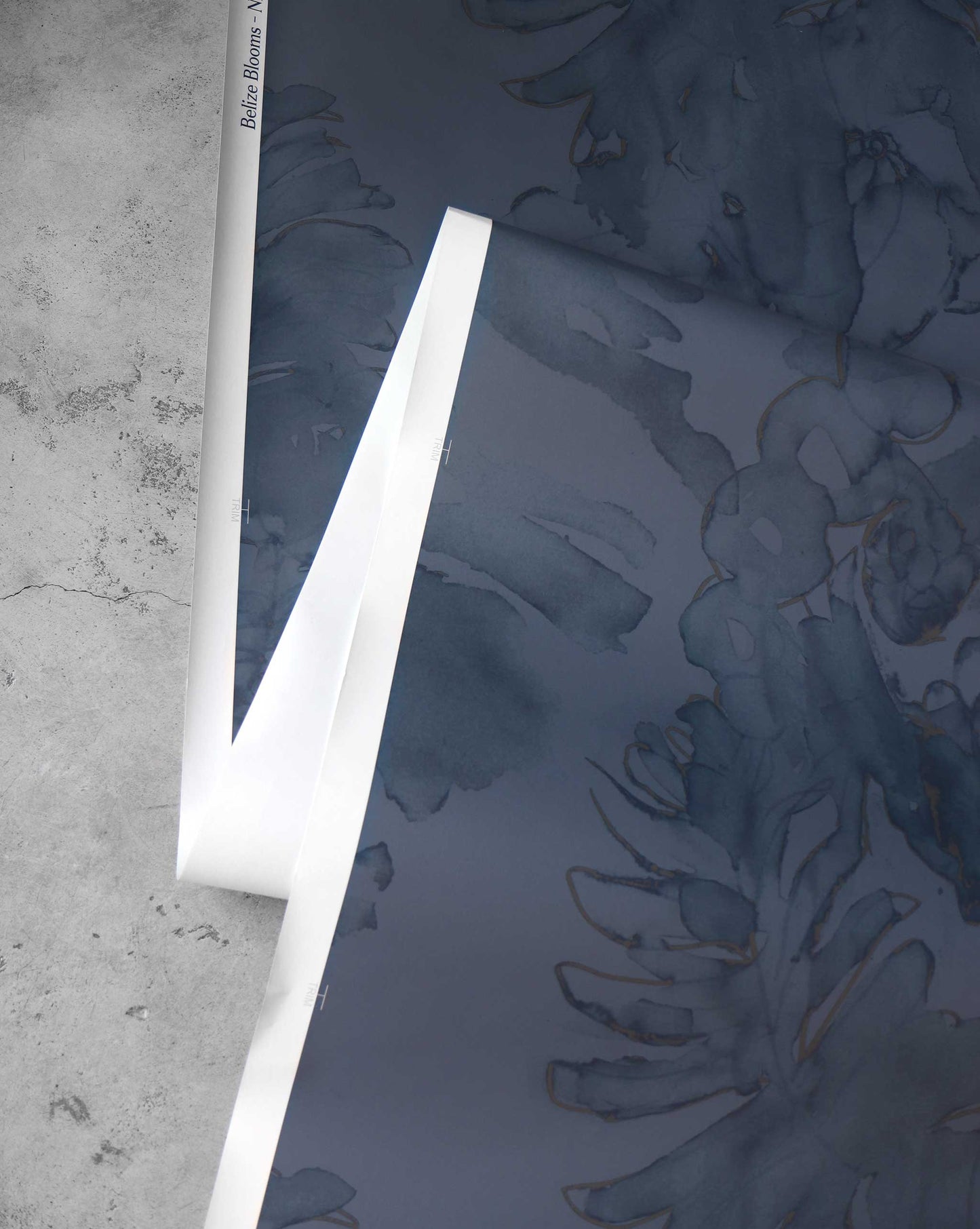 A dark blue Belize Blooms Wallpaper Mural||Night Fog with a floral pattern is partially unrolled on a grey surface, showcasing its tropical abundance and luxury mural vibes.