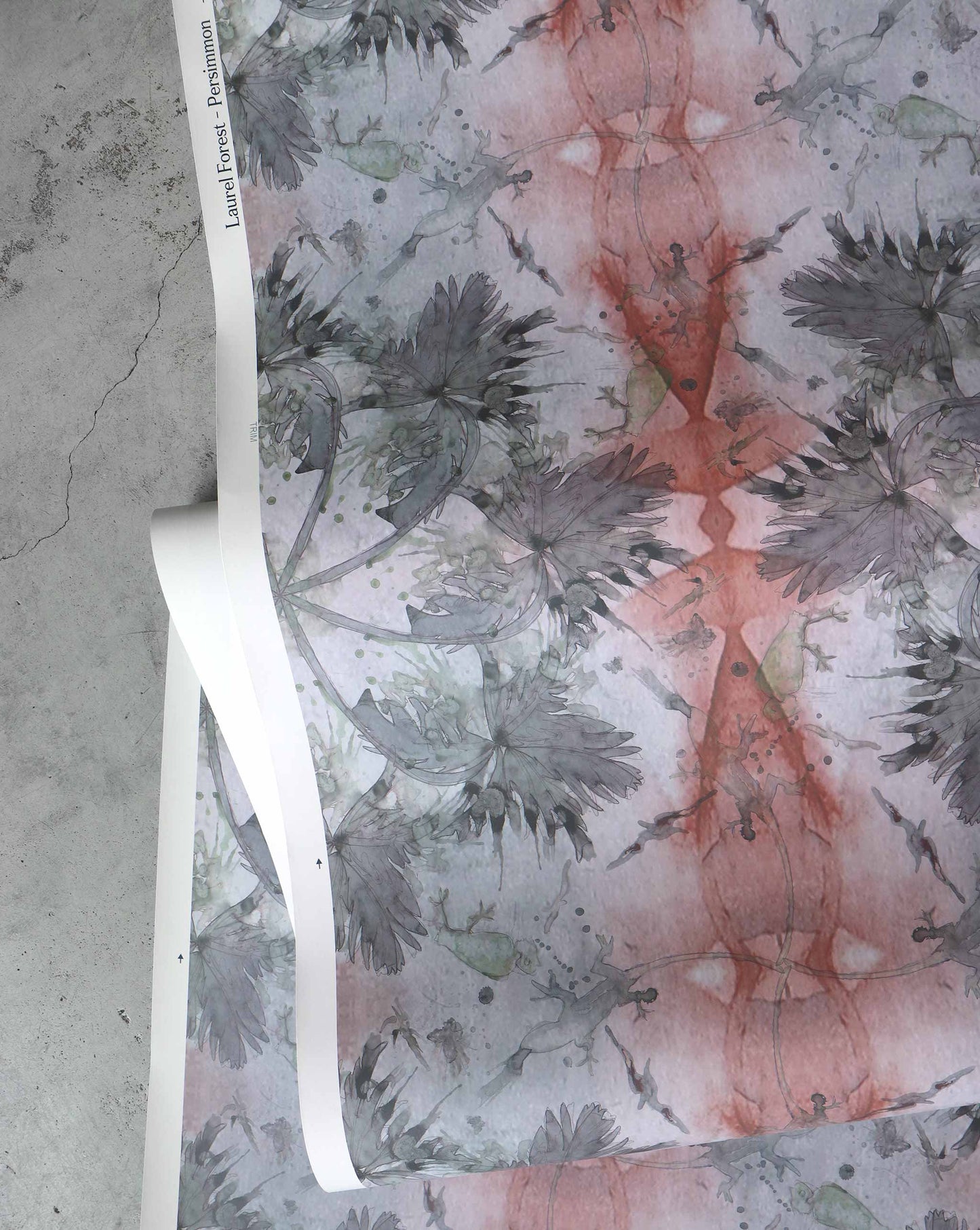 Rolled-out sheets of Laurel Forest Wallpaper||Persimmon with an abstract pattern featuring dark green and red hues lie on a gray surface, evoking a tropical atmosphere.