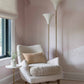 A cozy corner boasts a plush armchair, a small table with a vase of flowers, and two elegant floor lamps. The room is softly lit, featuring pastel pink walls adorned with Mod Wallpaper Mural||Shell that adds to the serene ambiance.