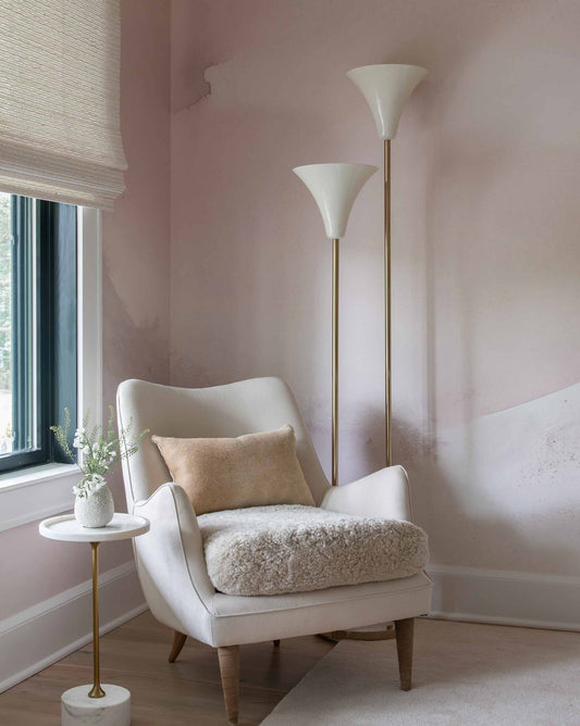 A cozy corner boasts a plush armchair, a small table with a vase of flowers, and two elegant floor lamps. The room is softly lit, featuring pastel pink walls adorned with Mod Wallpaper Mural||Shell that adds to the serene ambiance.