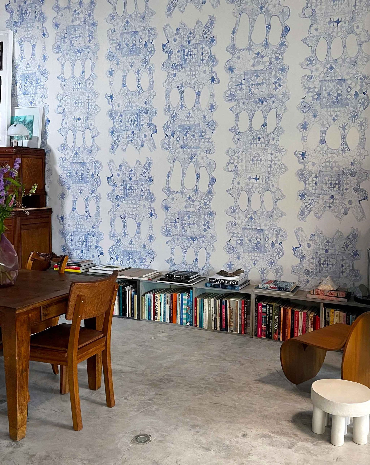 A room with Nonna's Lace Wallpaper in Fresco Blue, a wooden table and chair, a low bookshelf filled with books, and modern sculptural stools on a concrete floor.