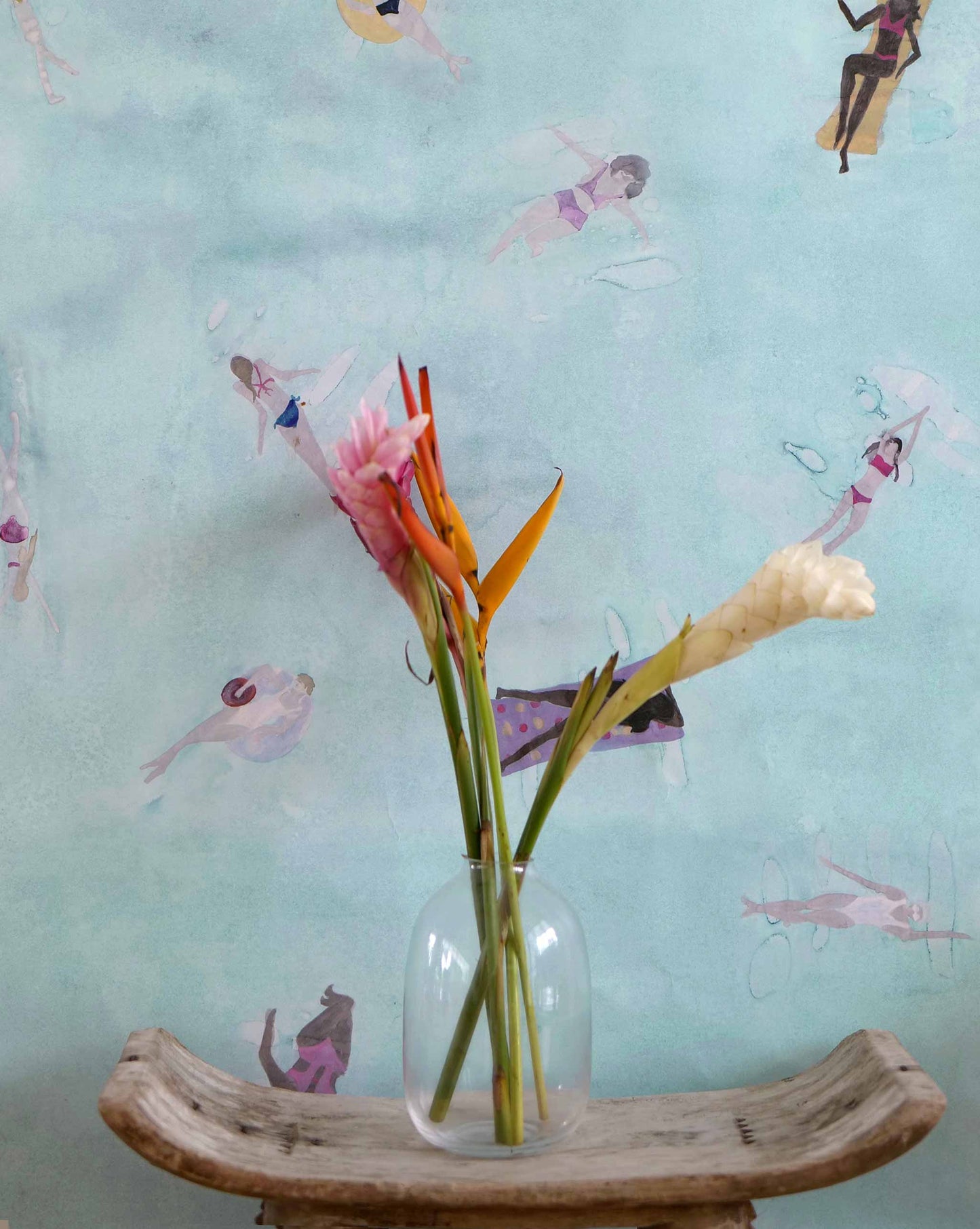 Eskayel's Swim wallpaper in the colorway Sea in light blue with waterborne female figures installed as an accent wall with a vase of flowers and a wooden stool.