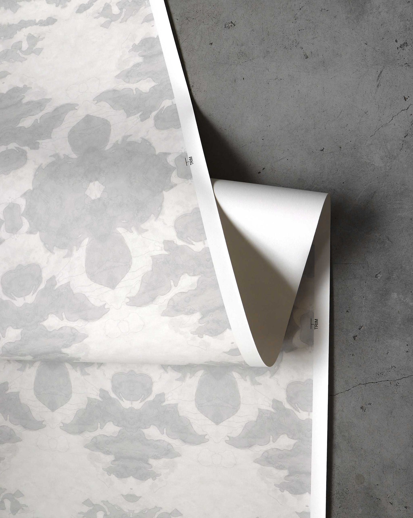 A piece of The Dance Wallpaper||Cloud from the Lora Collection, featuring a grey and white floral pattern with abstract elements, is partially unrolled on a grey concrete surface.