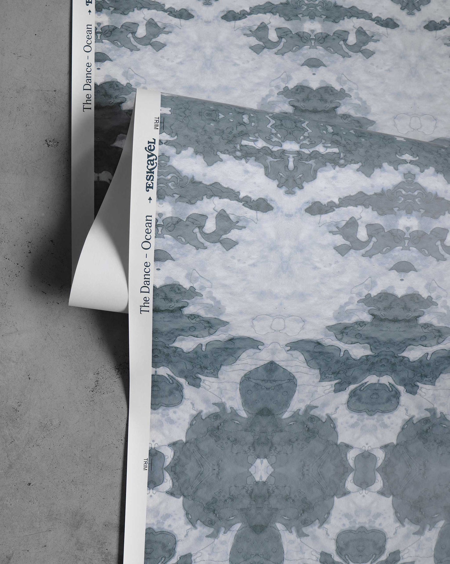 A partially unrolled sheet of luxury wallpaper with blue tones and gray abstract designs, labeled "The Dance Wallpaper||Ocean," resting on a concrete surface.