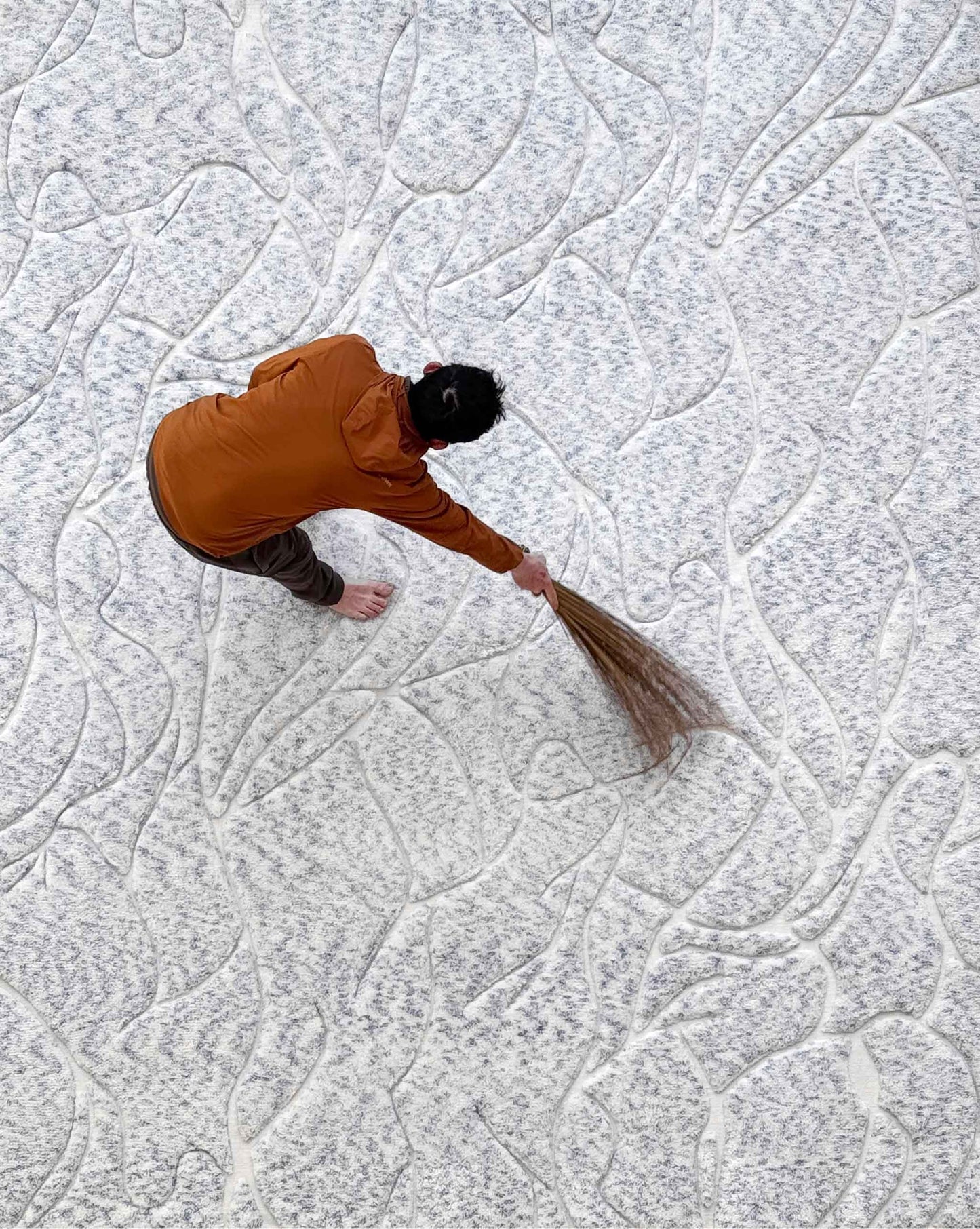 A person in an orange jacket sweeps a Hibiscus Lily Hand Knotted Rug in Sky from a top-down viewpoint.