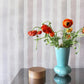 A vase of orange flowers on a table with a Gradient Stripe Grasscloth Pink Island behind it