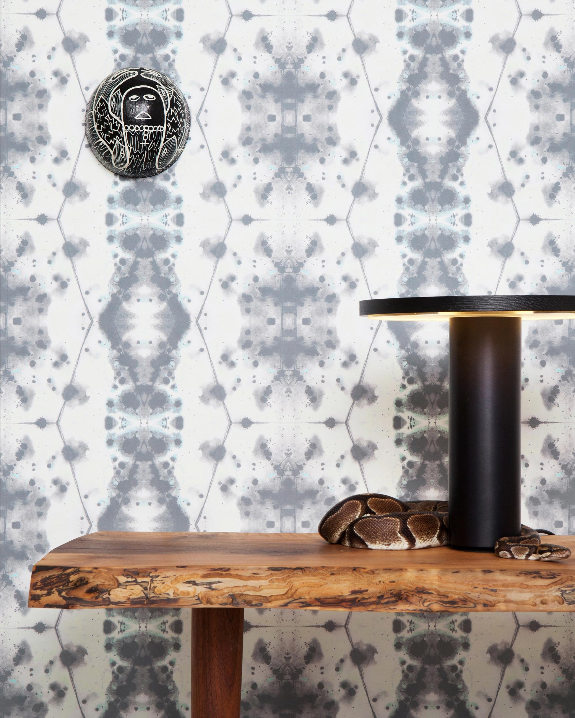 A table with a lamp and a clock in front of it, featuring the Hive Wallpaper in Waterstone colorway