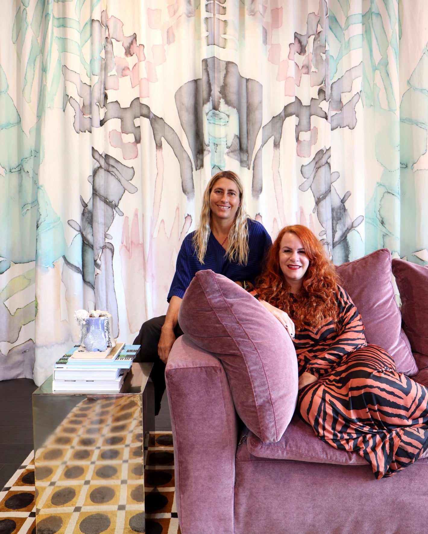Two women sitting on a purple couch in front of a colorful wall