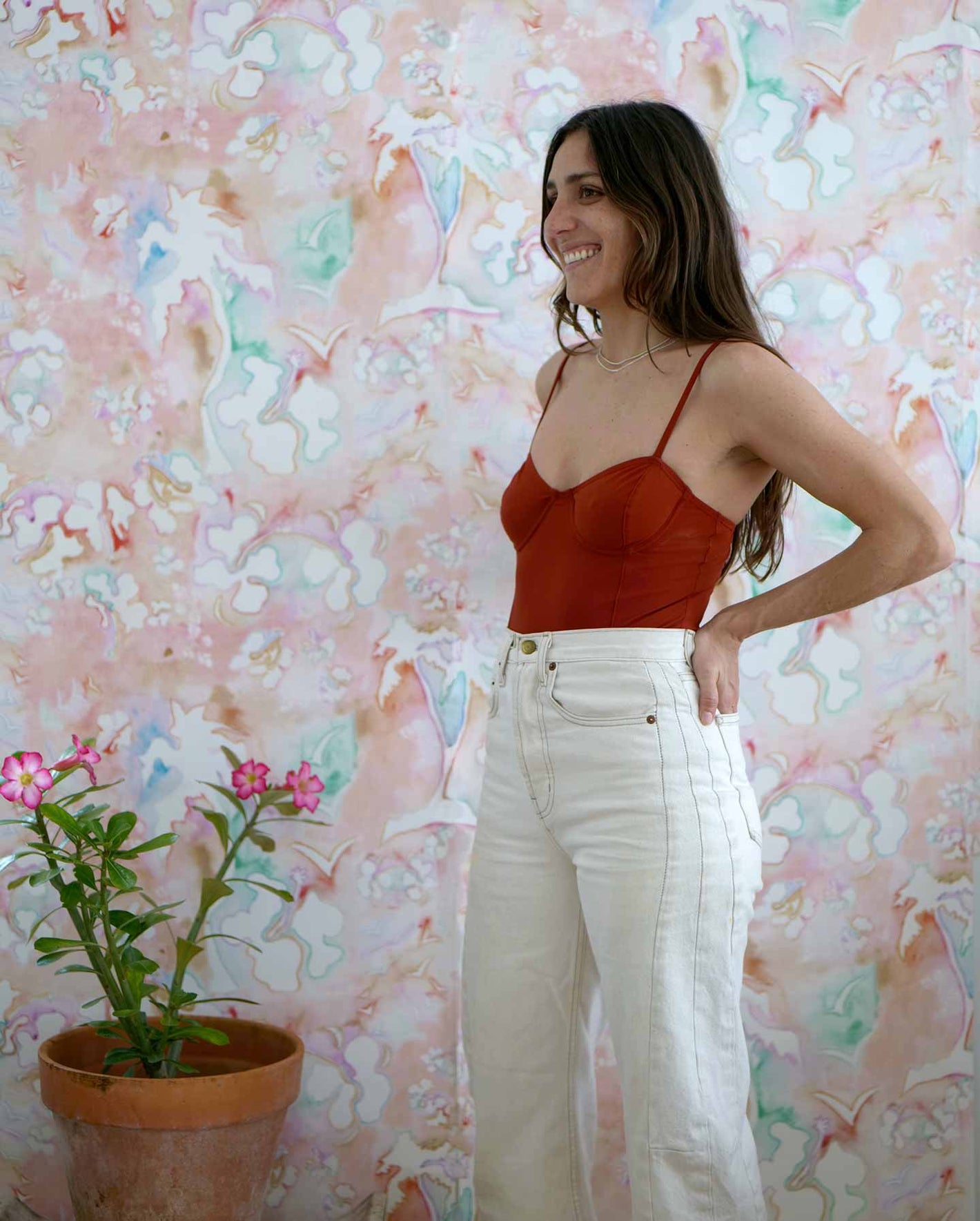 A woman posing in front of a red toned floral wallpaper with a vase of flowers.