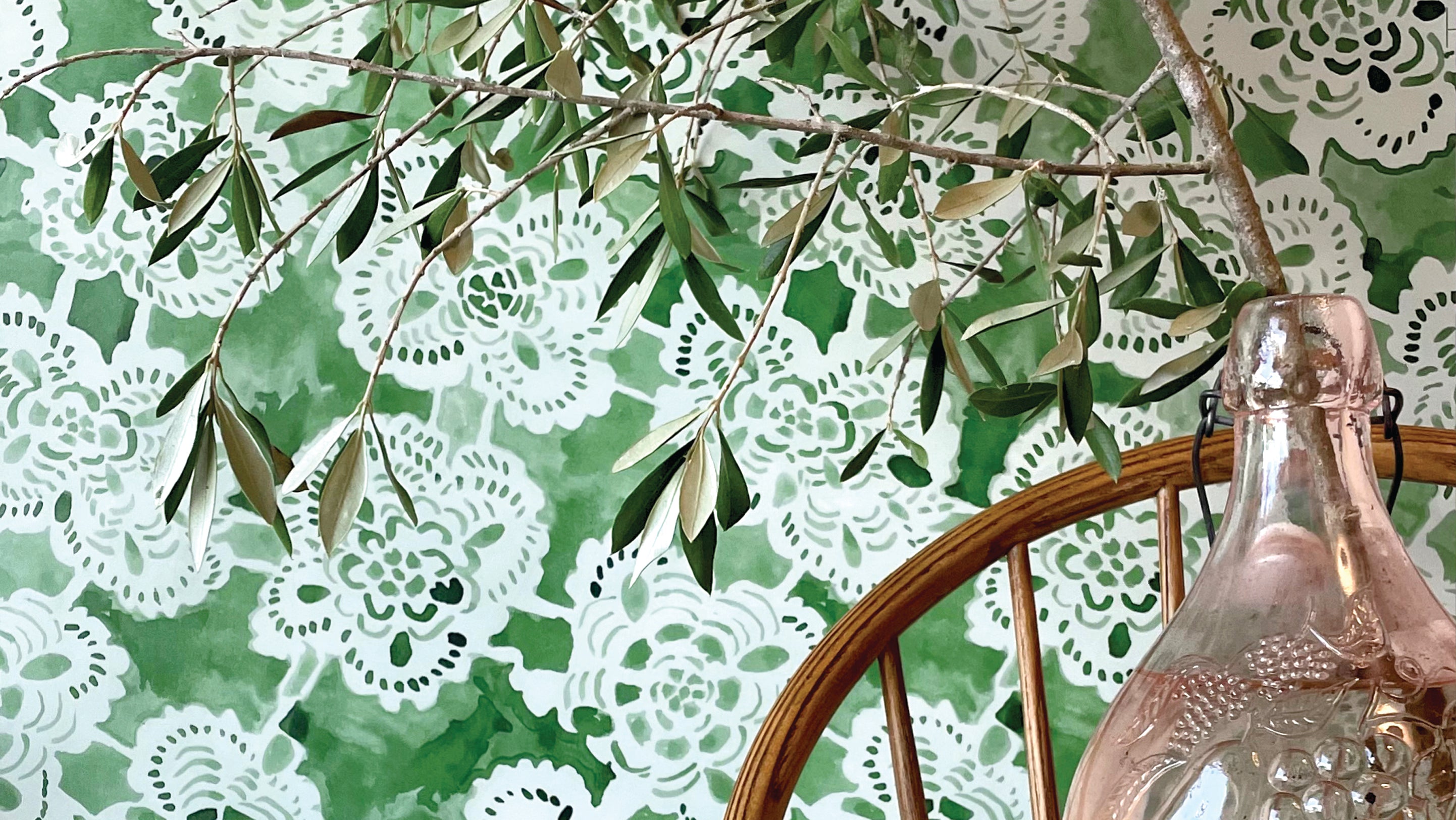 Clear glass bottle in front of a green lace wallpaper with olive branches.