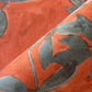 Close-up of Custom Grasscloth in orange, featuring a folded seam and a gray-green leaf pattern. The texture and weave are clearly visible, reminiscent of high-end custom designs.