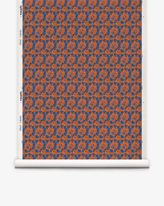 A roll of Alhelí Grasscloth in Lobelia, featuring a repeating pattern of blue floral sprays and dark thin stems on a textured, rust-orange background.