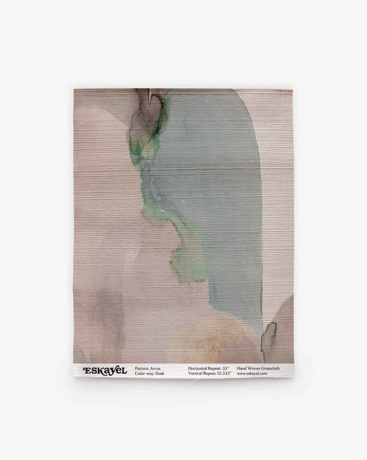 A fabric swatch showcasing abstract watercolor patterns in muted green, pink, and beige tones evokes the artistry of hand-painted designs. A label at the bottom lists the pattern as “Arcos Silk Wallcovering” in the color “Dusk,” recalling Eskayel’s elegant wallcovering aesthetic.