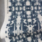 A piece of fabric featuring a blue and white abstract symmetrical pattern, reminiscent of Biami Grasscloth||Nila wallpaper, labeled “Biami Grasscloth||Nila” on the edge, partially folded on a gray surface.
