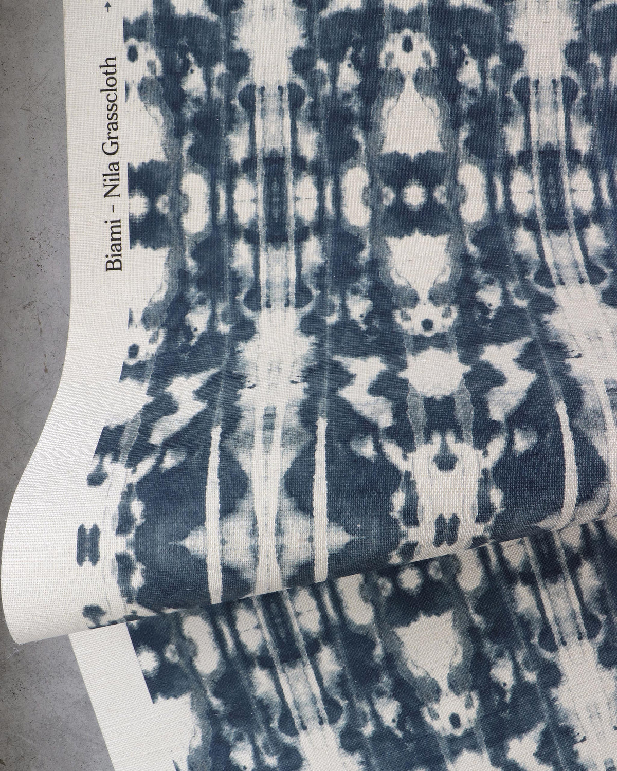 A piece of fabric featuring a blue and white abstract symmetrical pattern, reminiscent of Biami Grasscloth||Nila wallpaper, labeled “Biami Grasscloth||Nila” on the edge, partially folded on a gray surface.