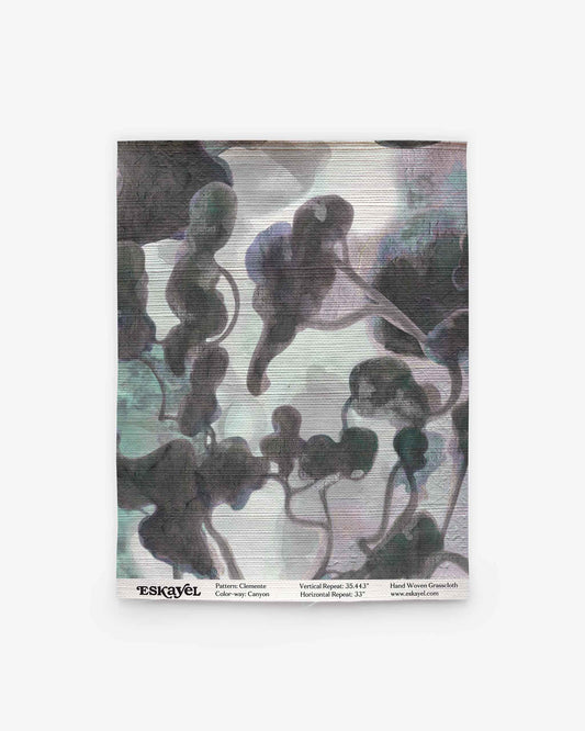 A square fabric swatch features an abstract, cloud-like watercolor pattern in muted gray, green, and purple tones on a light background, evoking the refined style of the Clemente Grasscloth||Canyon collection. Subtle text details and logo appear along the bottom edge, highlighting its inspiration from custom grasscloth wallpaper designs with abstracted tree motifs.
