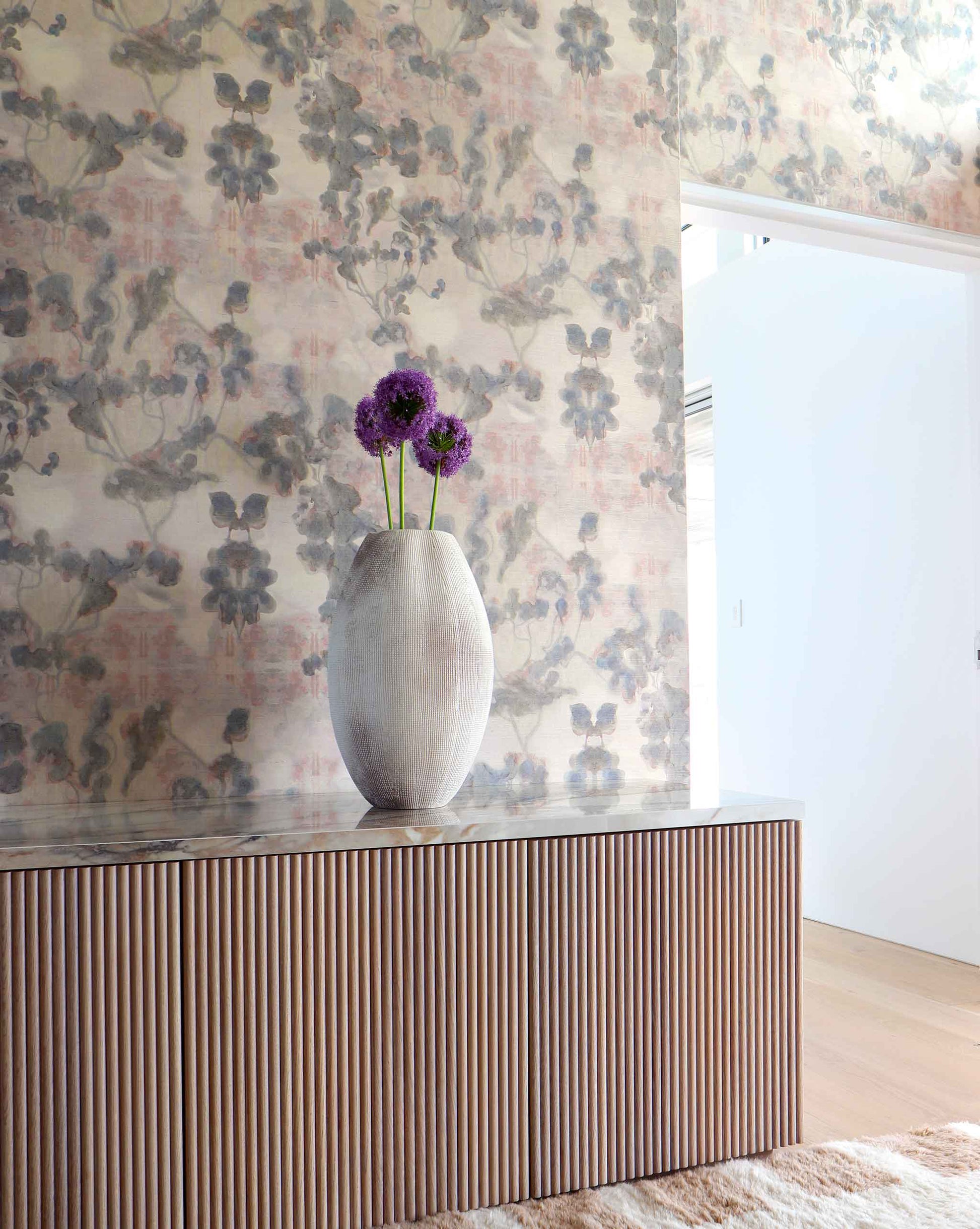 Vase with purple flowers on a wooden credenza against Eskayel's Clemente grasscloth wallpaper in sol.