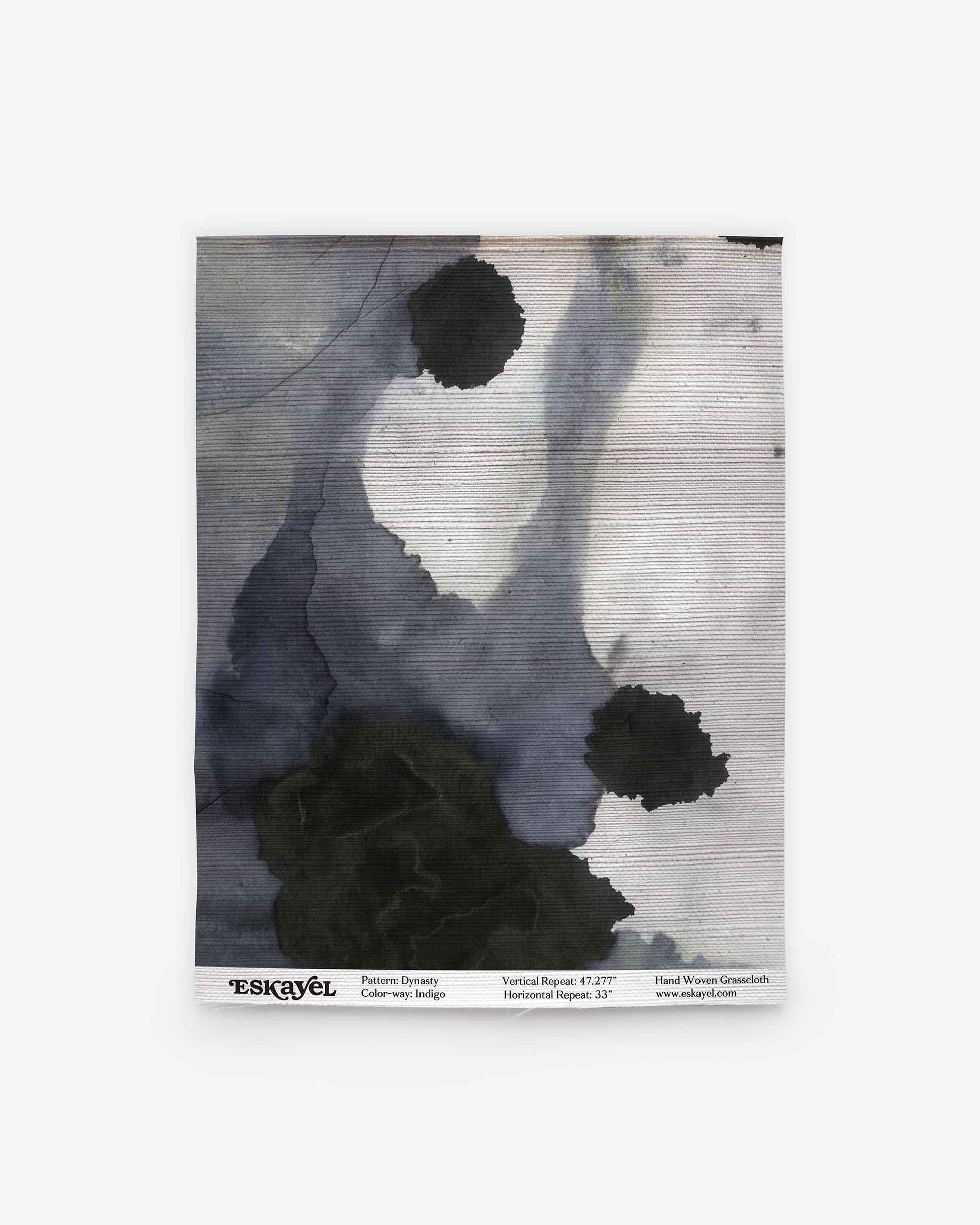 A rectangular textile sample featuring abstract black and gray watercolor-like stains, reminiscent of luxury wallpaper designs, is labeled “ESKAYEL Dynasty Grasscloth||Indigo” along the bottom edge.