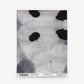 A square hand-woven textile, the Galileo Glass Grasscloth||Indigo features black irregular spots and gray shading on a light background, evocative of abstract-patterned wallpaper. The branding and pattern details are printed along the bottom edge.