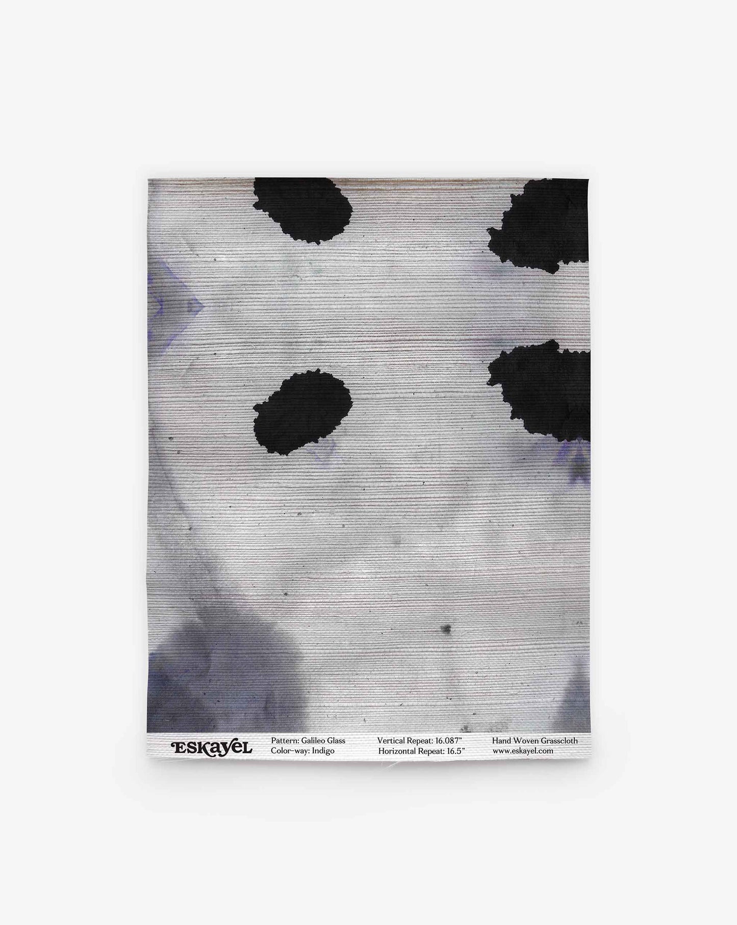 A square hand-woven textile, the Galileo Glass Grasscloth||Indigo features black irregular spots and gray shading on a light background, evocative of abstract-patterned wallpaper. The branding and pattern details are printed along the bottom edge.
