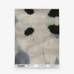 A folded textile featuring the Galileo Glass Grasscloth in the Shale colorway, showcasing an abstract black spot pattern on a light gray background, displayed against a white surface—evoking high-end wallpaper aesthetics. Text at the bottom notes the pattern, color (Shale), size, and brand details.