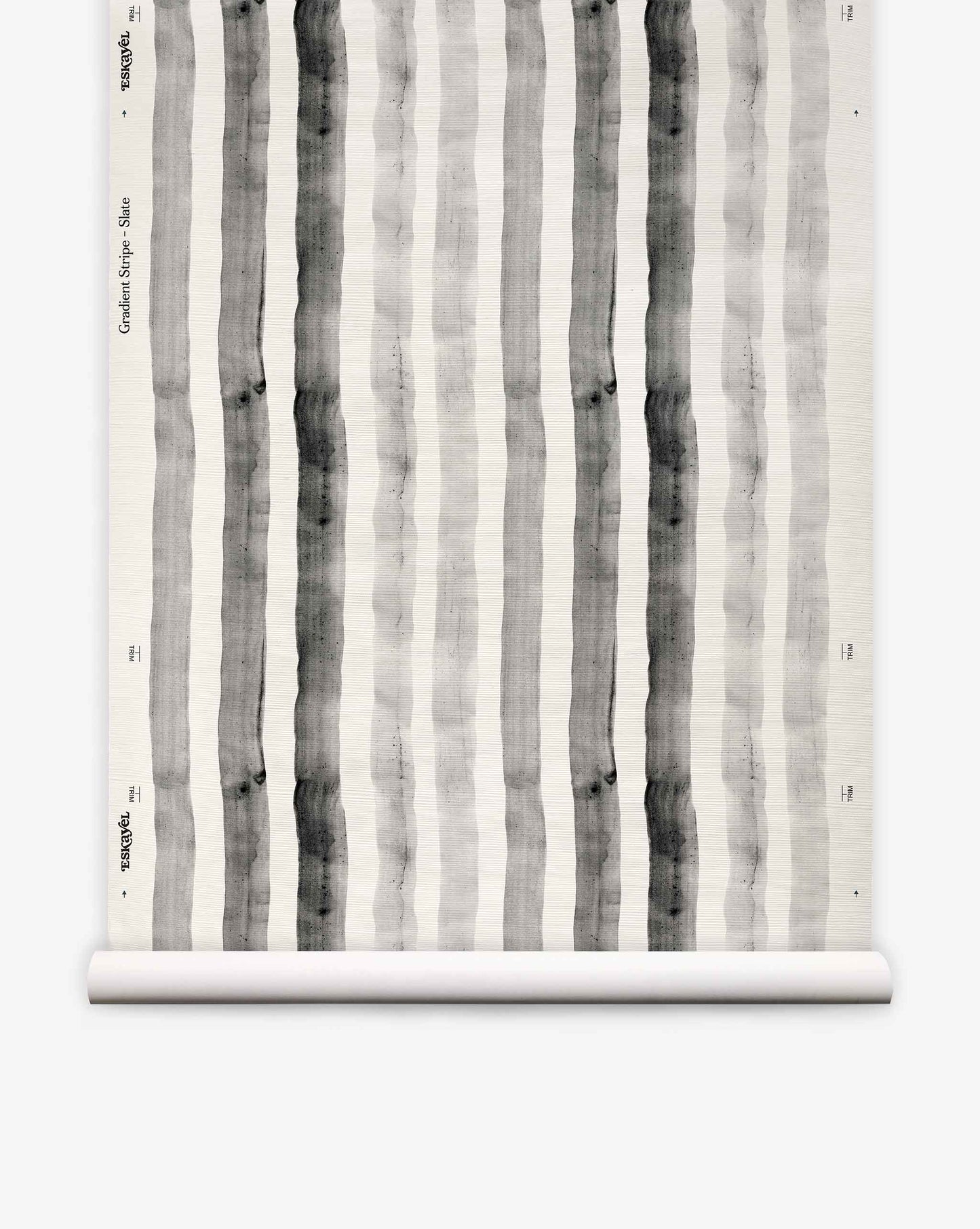 A roll of “Gradient Stripe Grasscloth||Slate” wallpaper, featuring vertical black and gray watercolor stripes on a white background, is partially unrolled and displayed against a plain surface, evoking a subtle nautical vibe with its watercolor-inspired design.