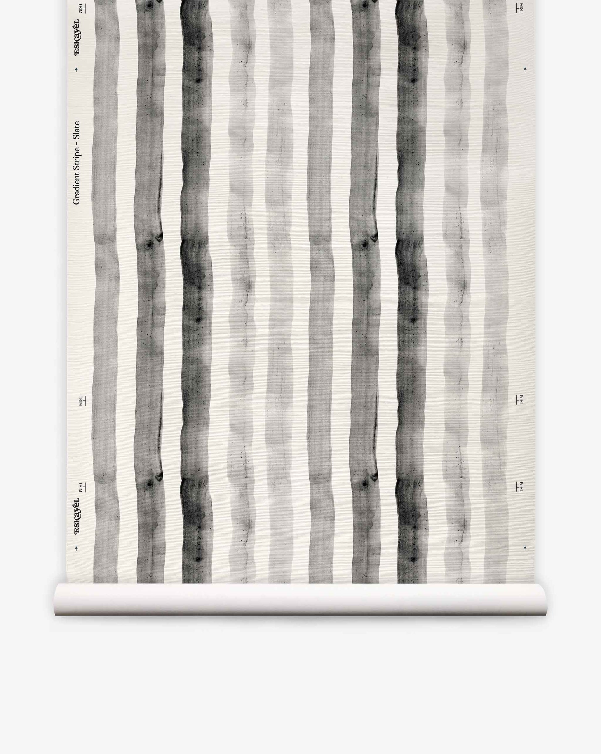 A roll of “Gradient Stripe Grasscloth||Slate” wallpaper, featuring vertical black and gray watercolor stripes on a white background, is partially unrolled and displayed against a plain surface, evoking a subtle nautical vibe with its watercolor-inspired design.