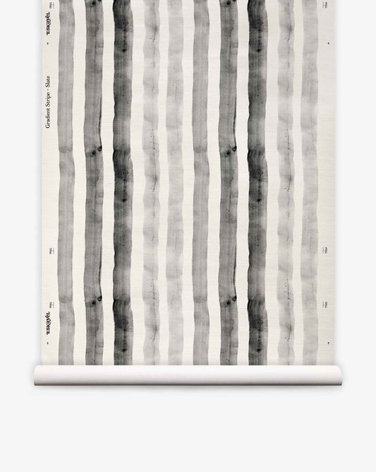 A roll of “Gradient Stripe Grasscloth||Slate” wallpaper, featuring vertical black and gray watercolor stripes on a white background, is partially unrolled and displayed against a plain surface, evoking a subtle nautical vibe with its watercolor-inspired design.