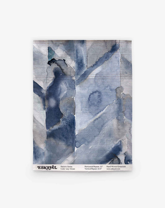 Abstract blue and gray watercolor textile swatch with overlapping geometric shapes evokes hand-woven wallpaper. Label: “Sorisa Grasscloth||Ocean” with pattern, color, and website details.