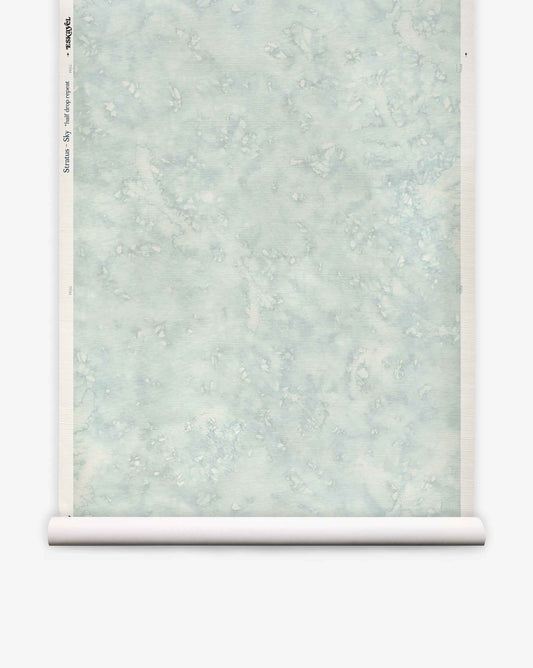 A partially unrolled roll of Stratus Grasscloth||Sky wallpaper, featuring a pale blue marbled design with subtle white and gray accents, is displayed against a white background.