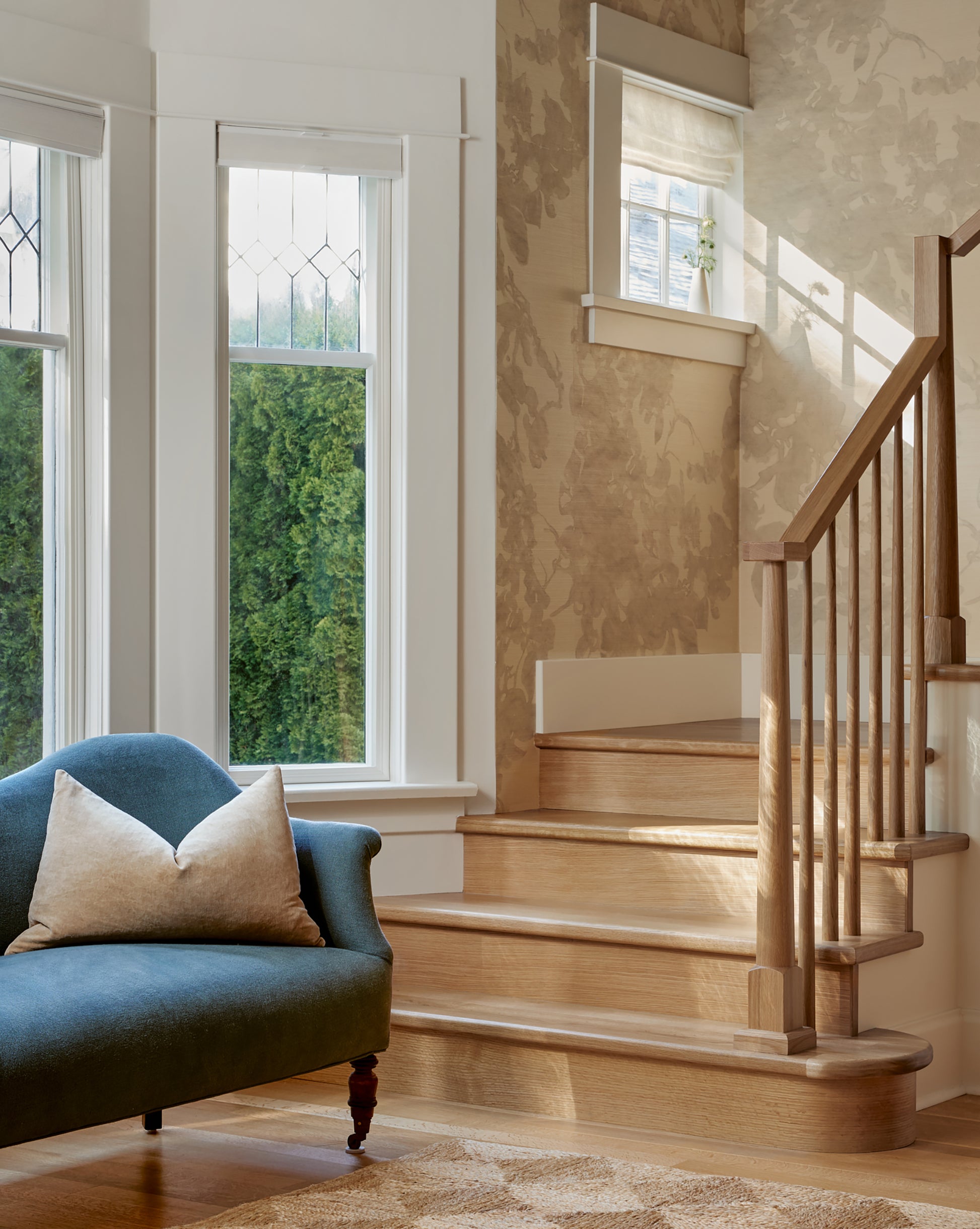 A wooden staircase by two large windows with white trim; a blue upholstered chair with a beige pillow sits in the foreground, accented by Up for Anything Grasscloth||Pearl wallcovering that adds texture to the space.