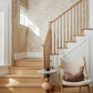 A wooden staircase with a light wood railing, Up for Anything Grasscloth||Pearl wallcovering, a modern side table, and a cushioned chair with a pillow in a bright interior.