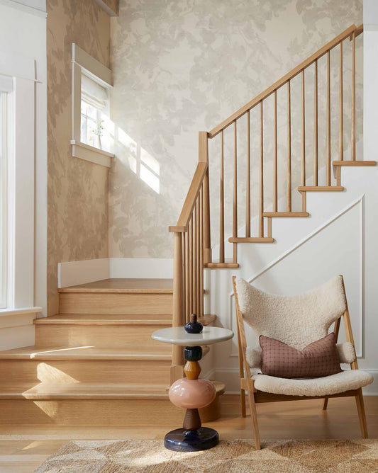 A wooden staircase with a light wood railing, Up for Anything Grasscloth||Pearl wallcovering, a modern side table, and a cushioned chair with a pillow in a bright interior.