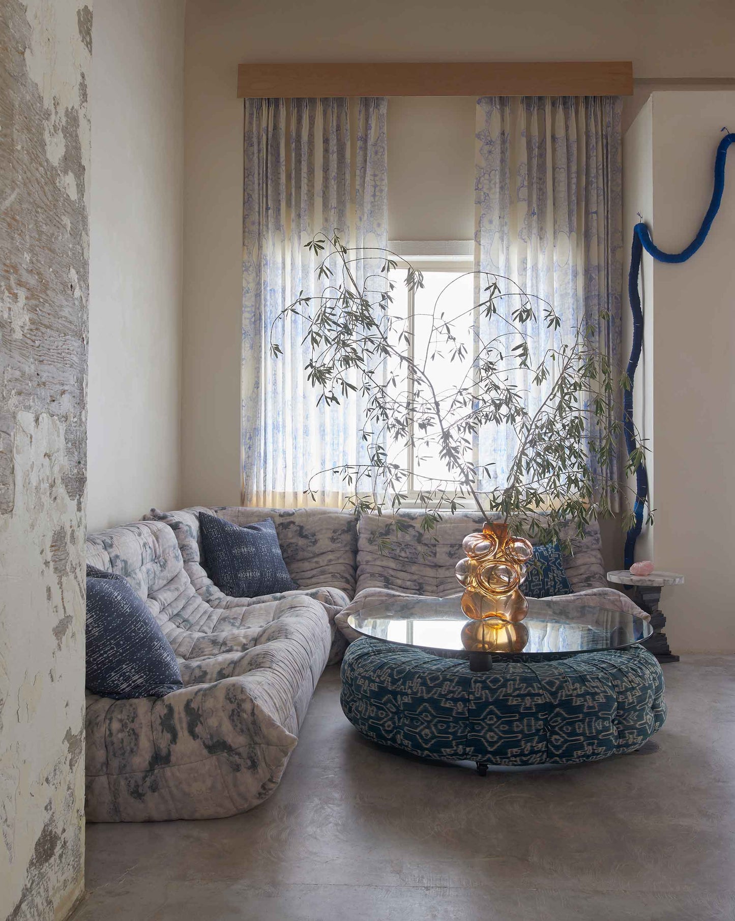 A cozy living room with a curved sofa, patterned pillows, round glass coffee table, potted plant centerpiece, and light filtering through Nonna's Lace Fabric in Fresco Blue.