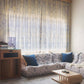 A cozy living room with Nonna's Lace Fabric in Fresco Blue curtains, a sectional sofa with cushions, a coffee table, and soft light streaming through the window.
