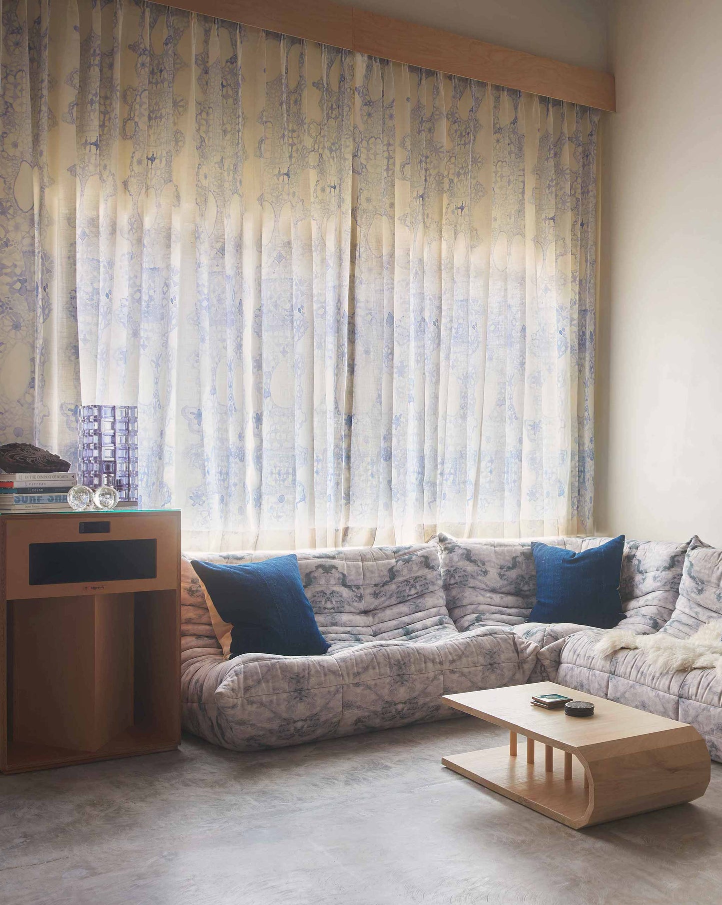 A cozy living room with Nonna's Lace Fabric in Fresco Blue curtains, a sectional sofa with cushions, a coffee table, and soft light streaming through the window.