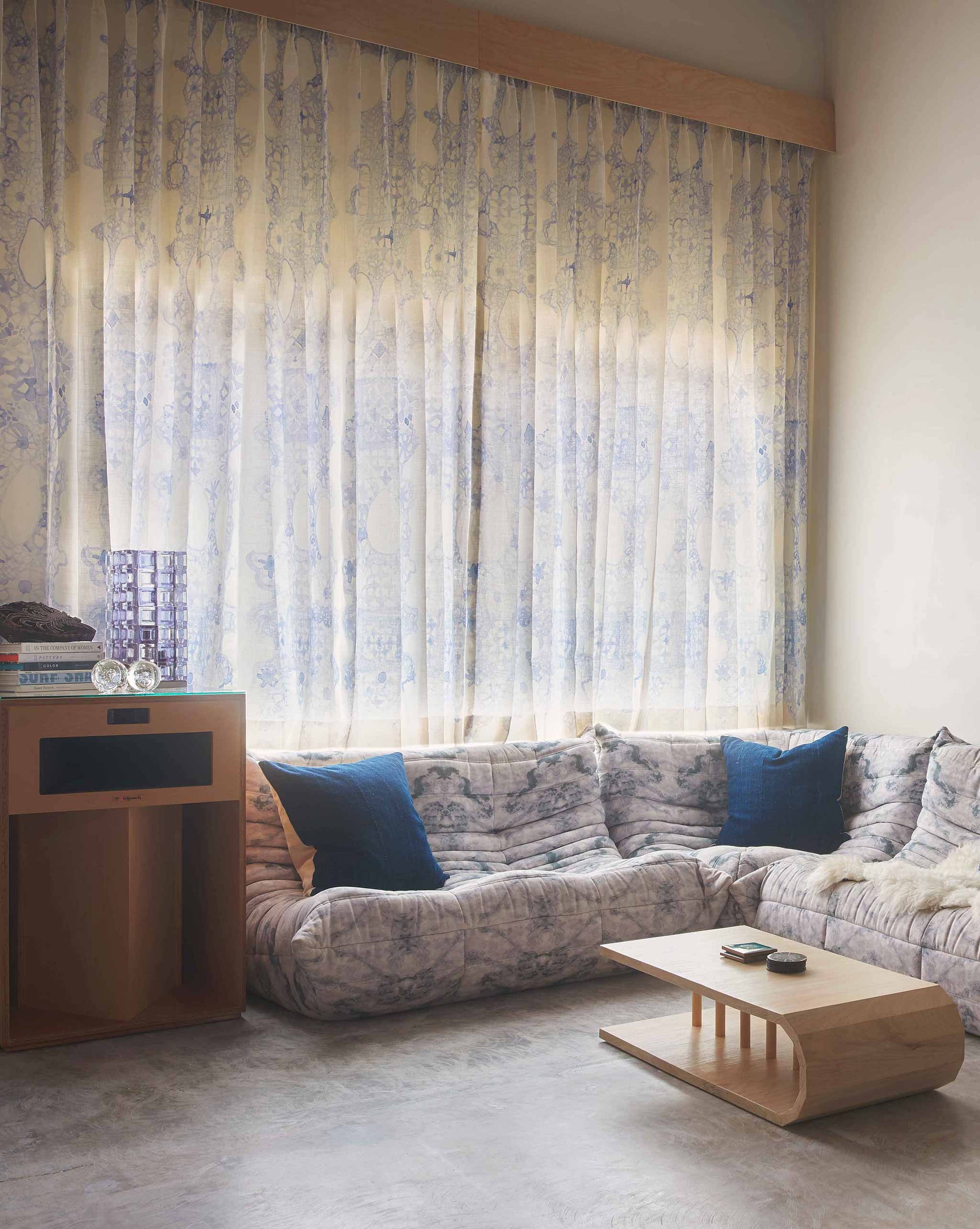 A cozy living room with Nonna's Lace Fabric in Fresco Blue curtains, a sectional sofa with cushions, a coffee table, and soft light streaming through the window.