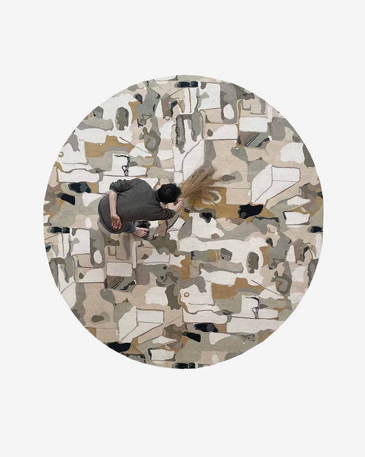 A person seen from above uses a broom on the Medina Hand Knotted Rug||Blanca CUSTOM, a circular, abstract-patterned handmade rug in neutral colors.
